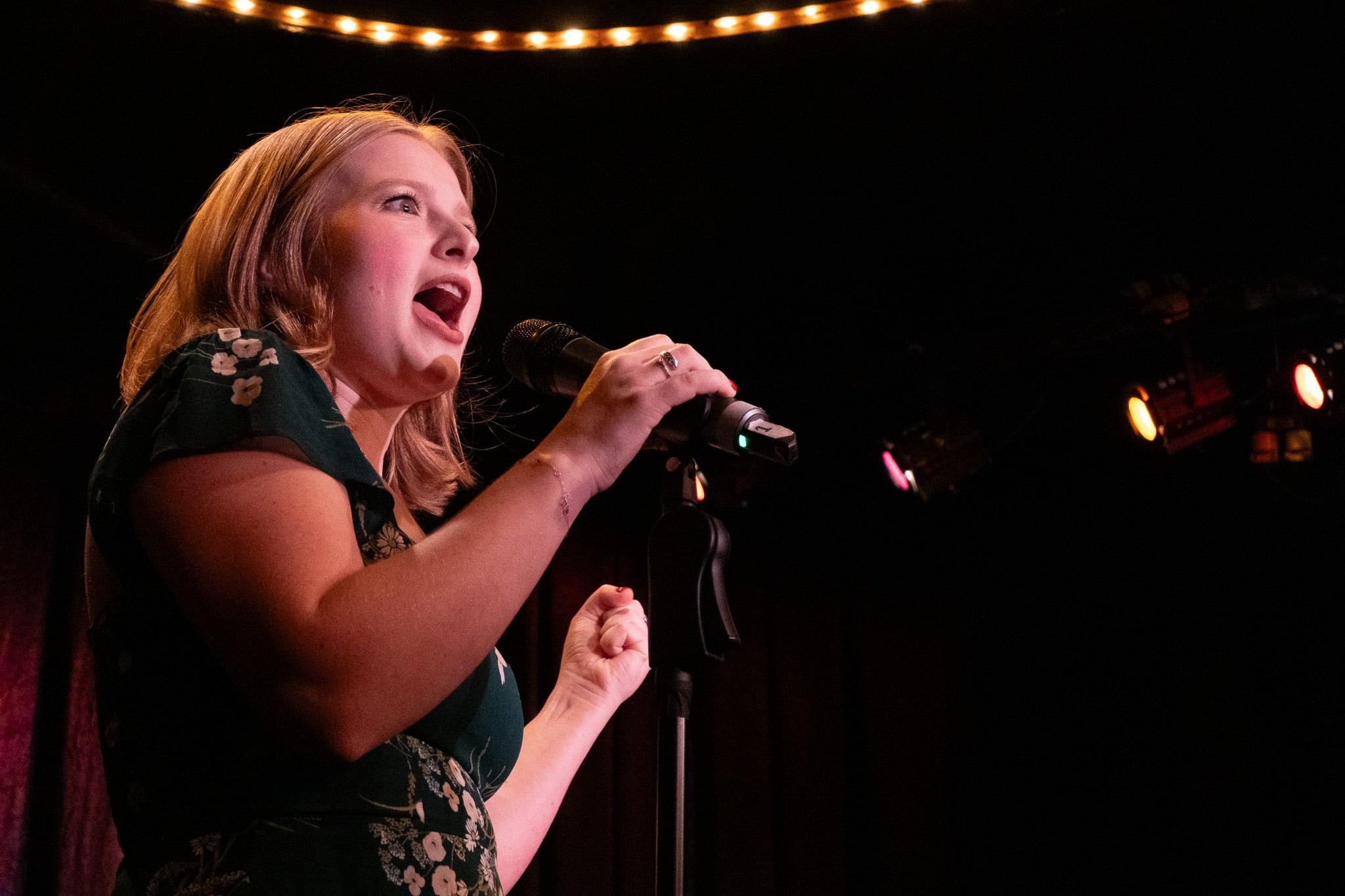 A close view of the singer belting into the microphone, string lights above.
