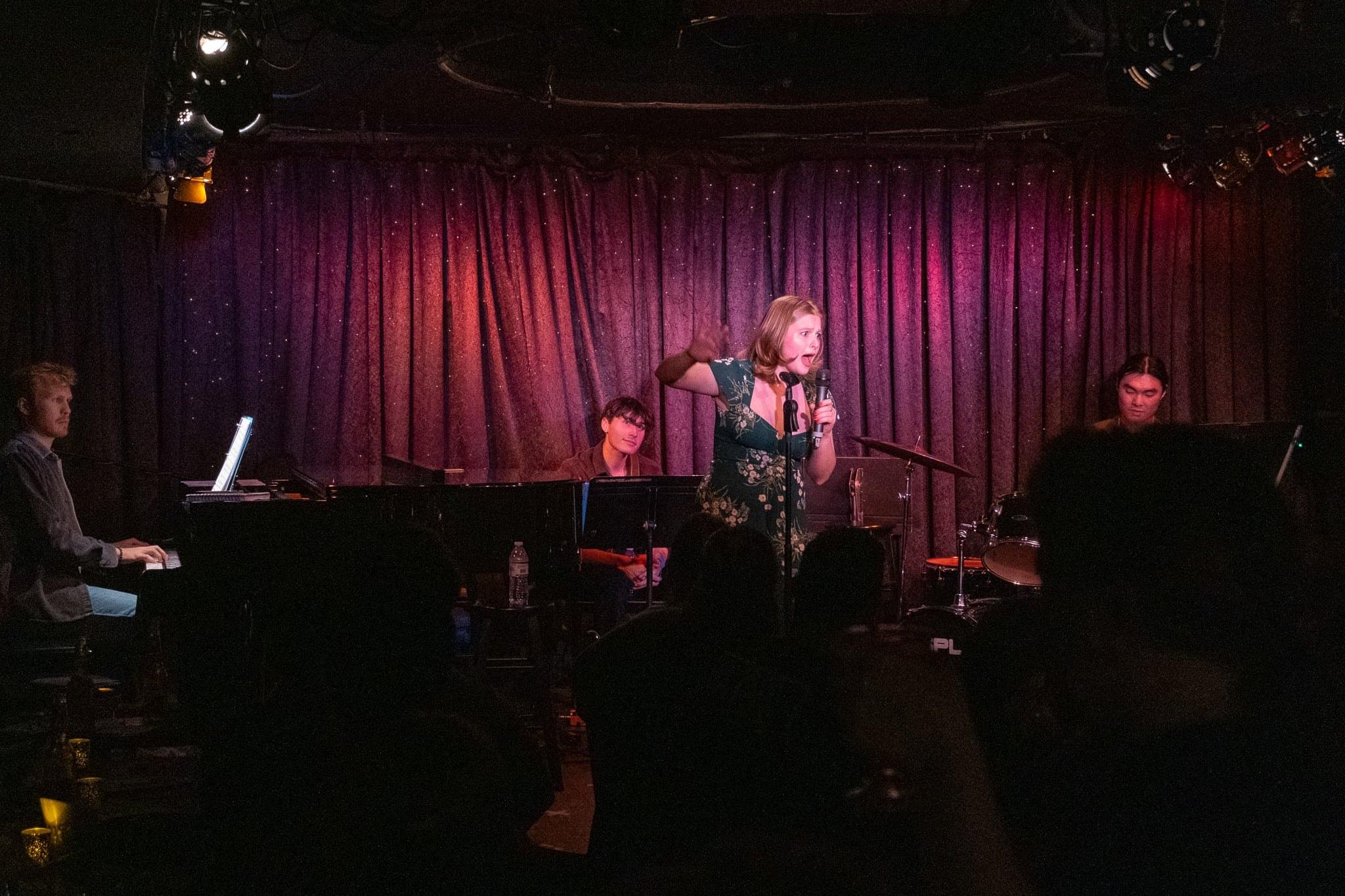 The whole stage from the back of the room: piano, band and singer under a purple curtain.