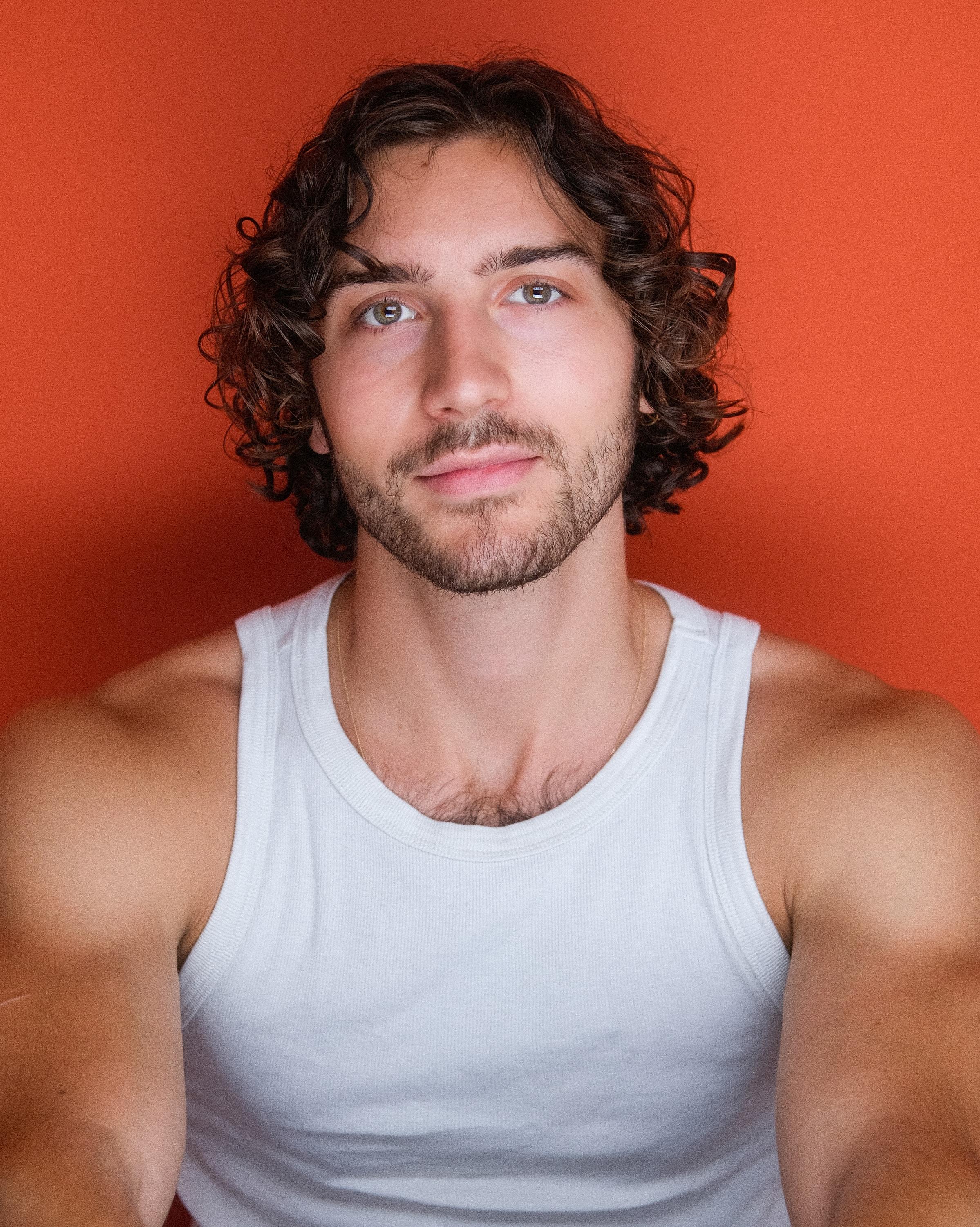 The same man in a white tank top, chin lifted, against a bright orange backdrop.
