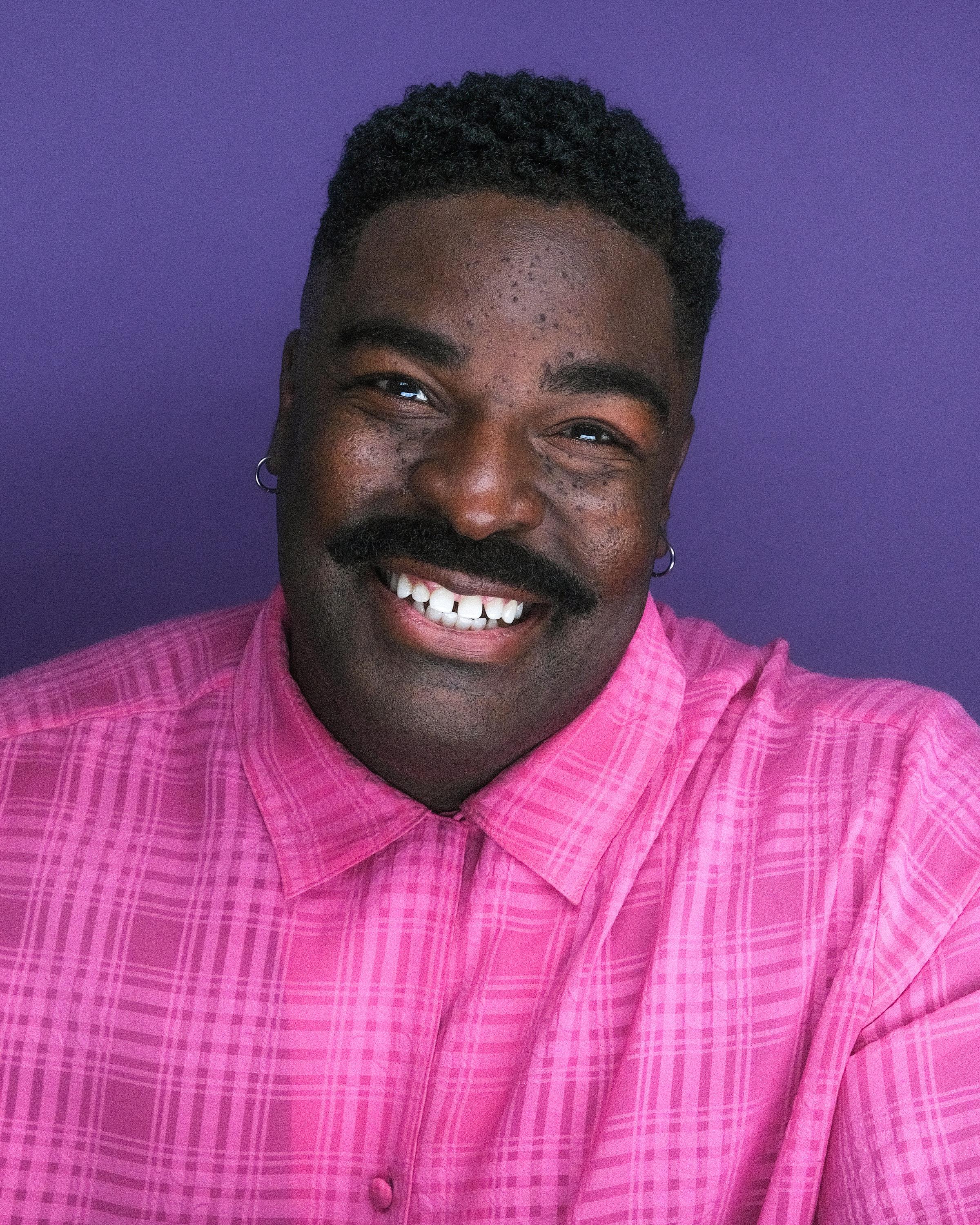The same man grinning in a pink plaid shirt against a violet backdrop.