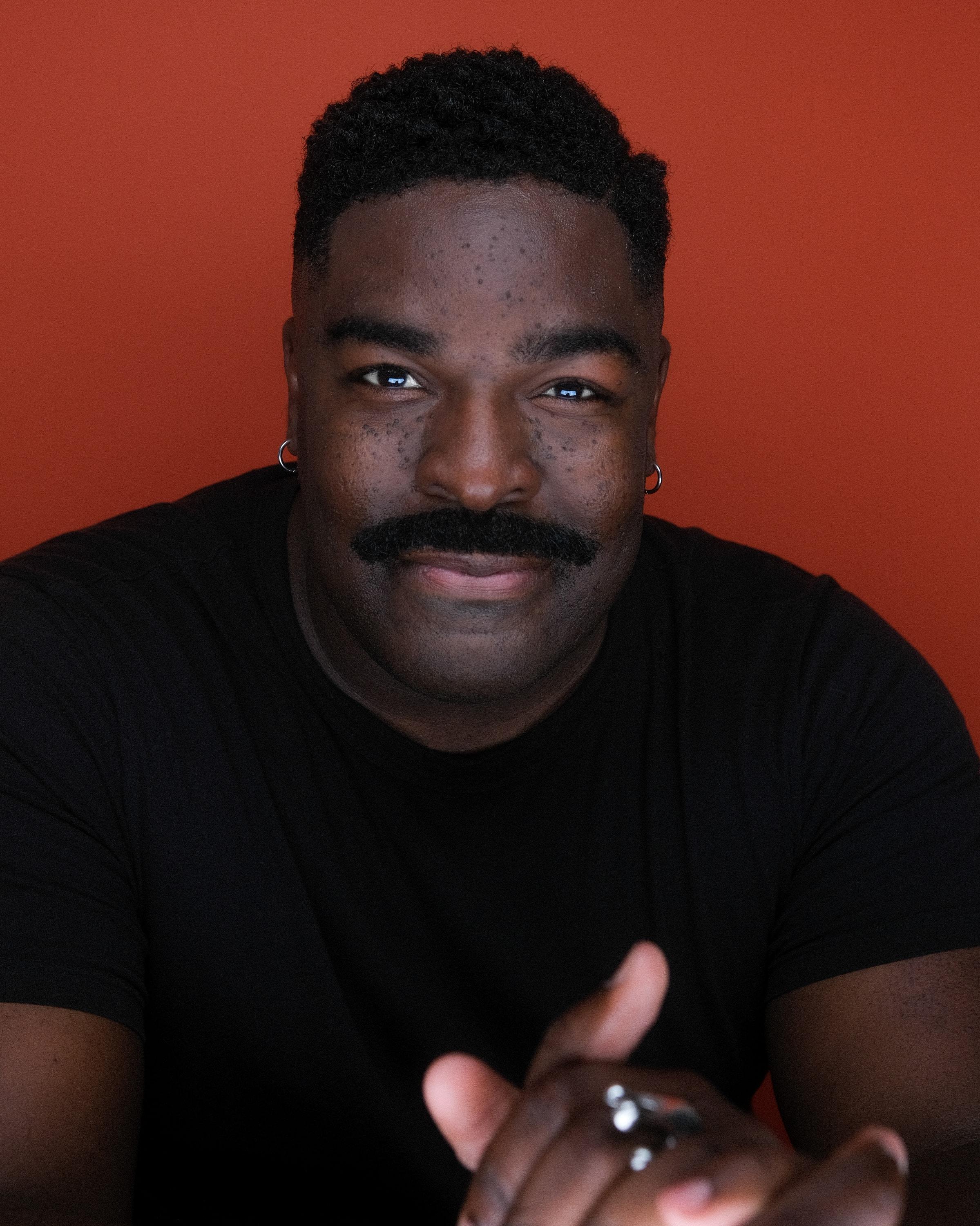 The same man in a black T-shirt, leaning toward the camera with a slight smile against a rust-red backdrop.