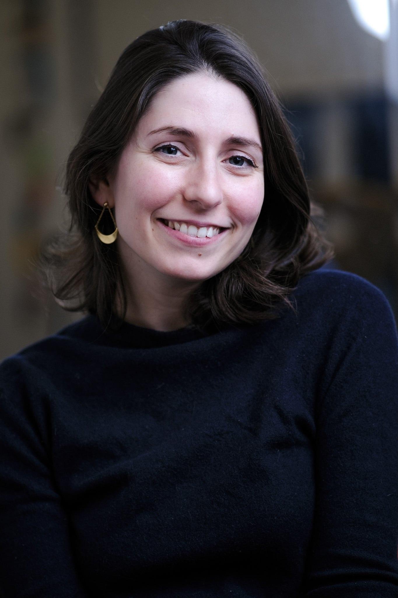 Woman with shoulder-length dark hair in a navy sweater, smiling, in a softly lit office.