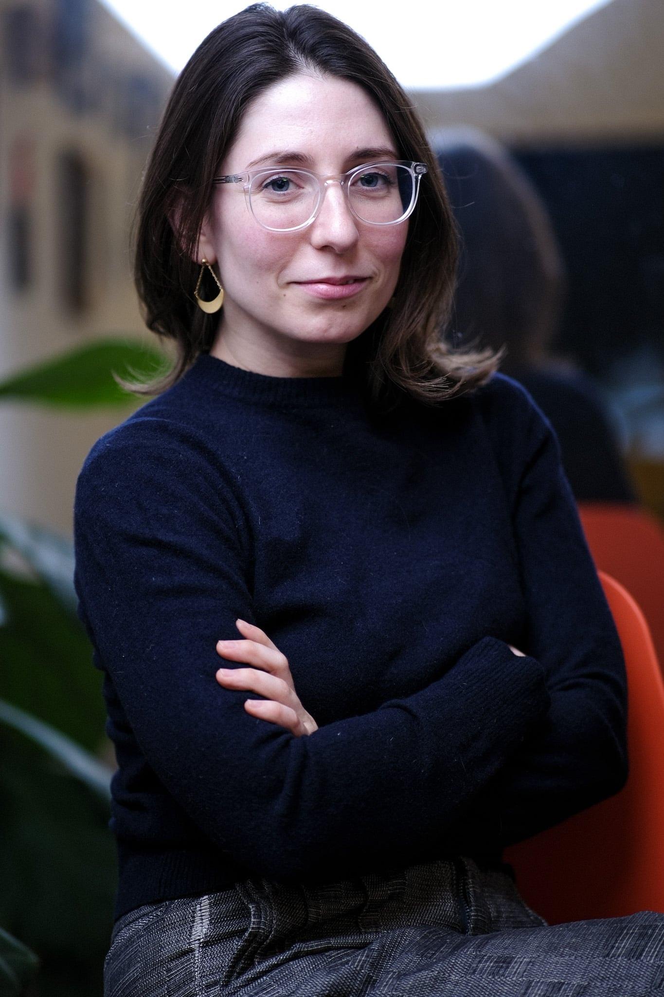 The same woman, arms crossed, in glasses, seated in the office.