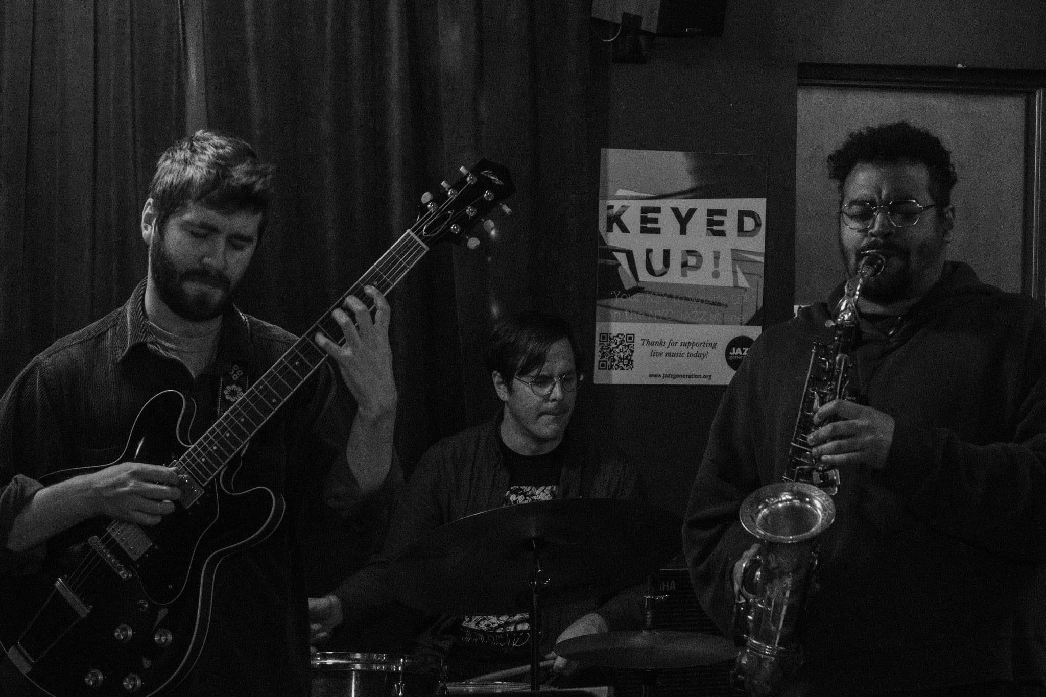 A guitarist and saxophonist playing side by side in a dim room, in black and white.