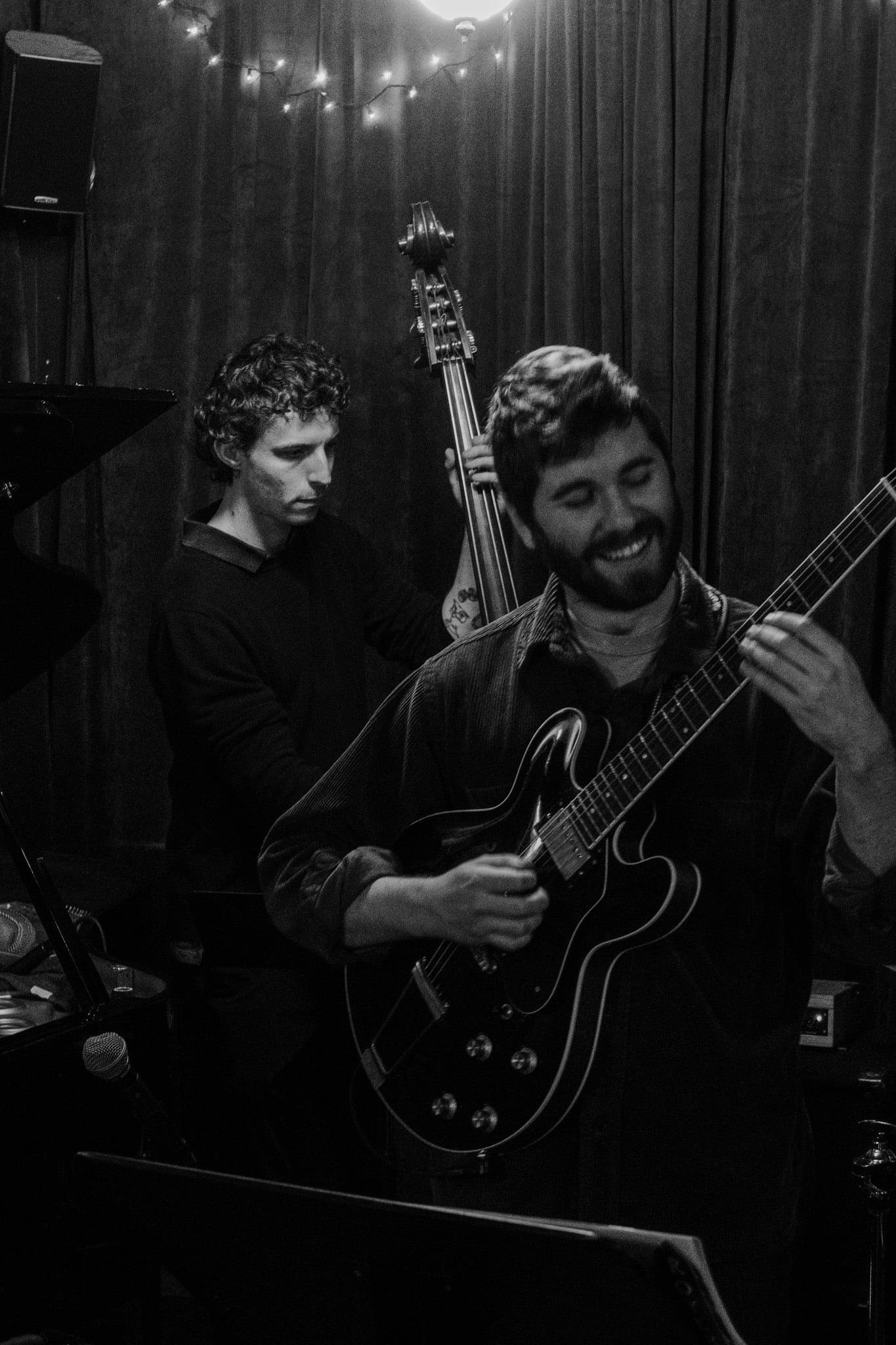 A guitarist smiling mid-solo, a bassist behind him, in black and white.
