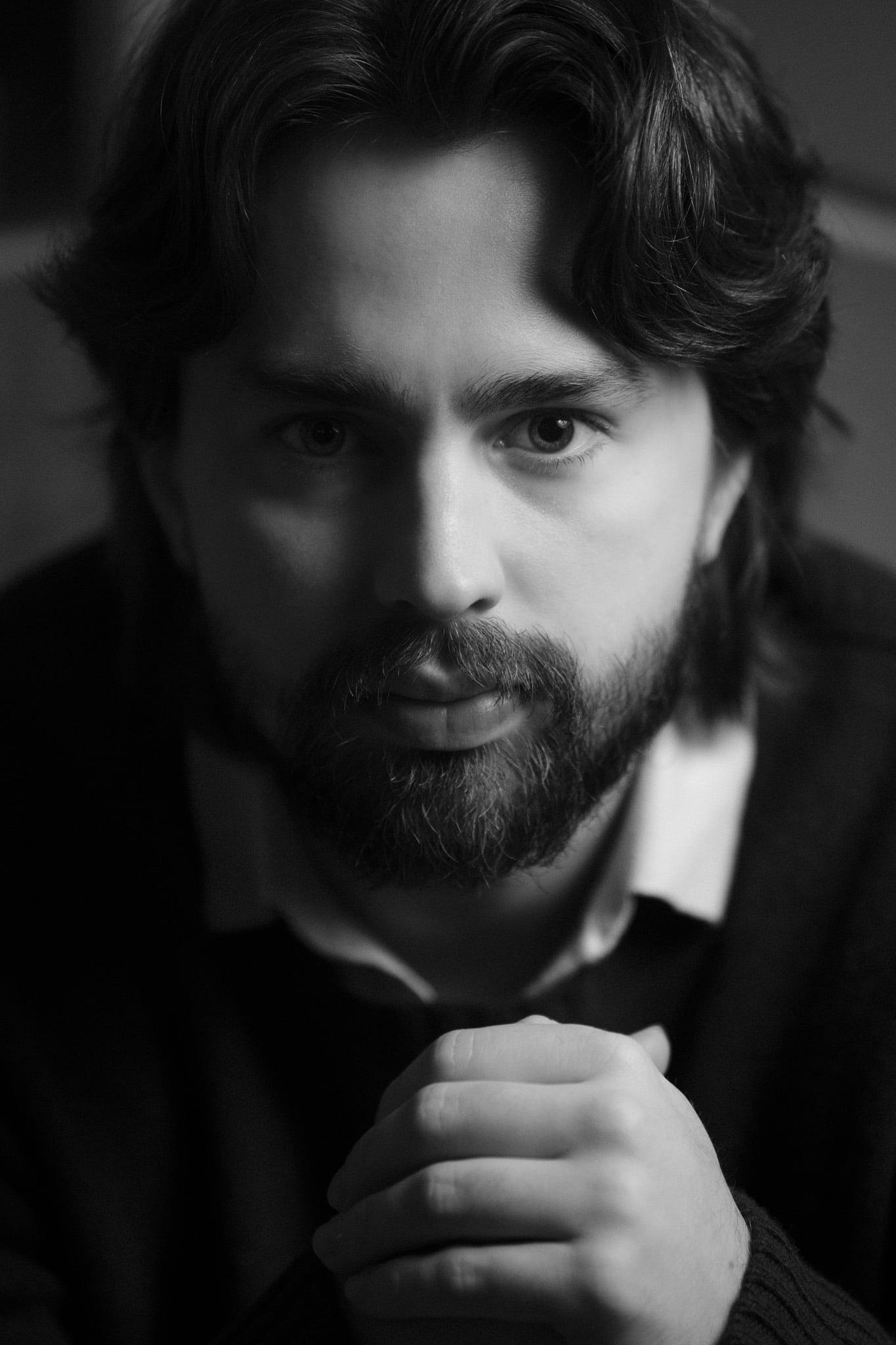 A bearded man with long hair, hands clasped under his chin, in black and white.