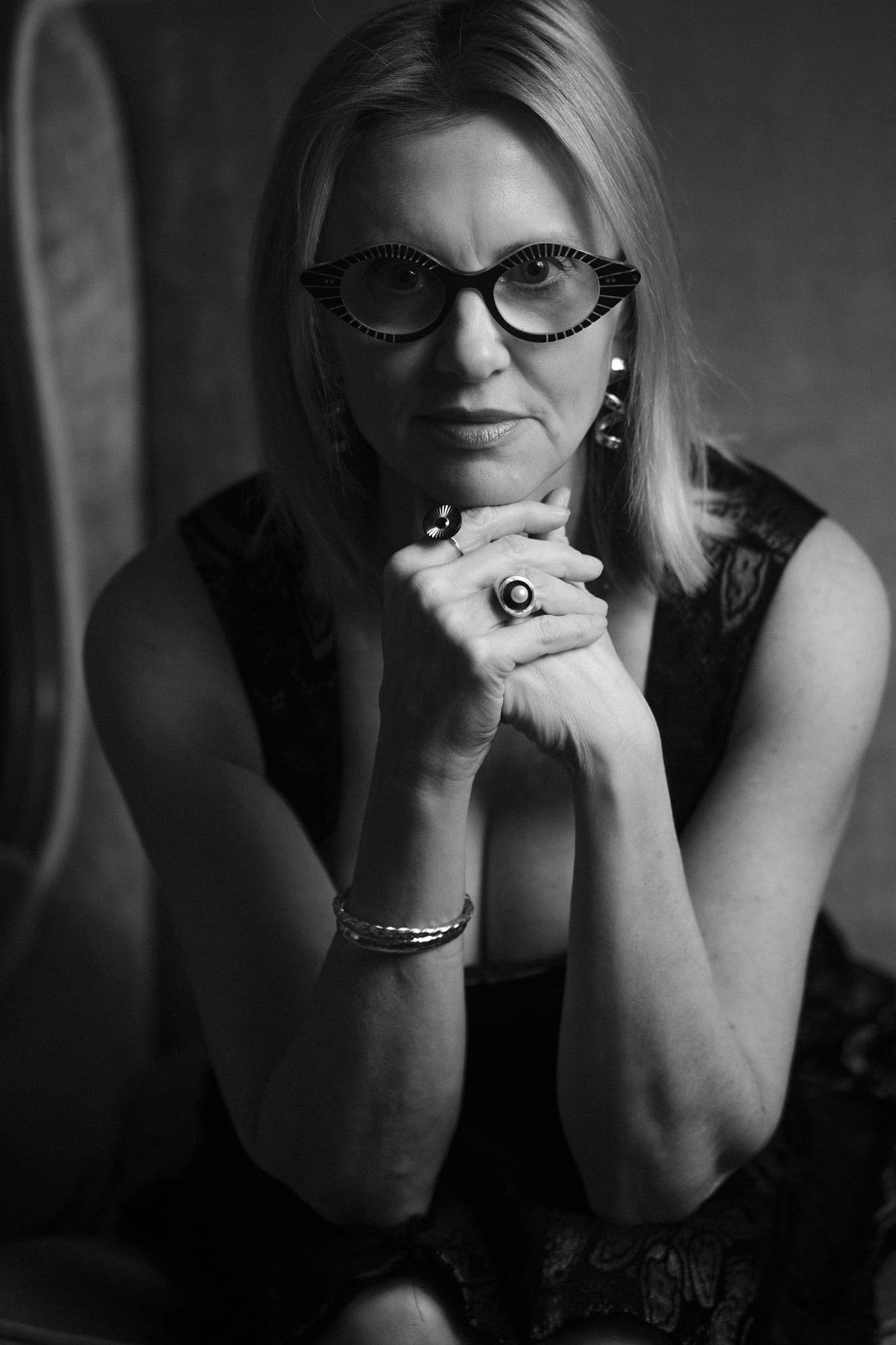 A woman with short blond hair and dark glasses, hands folded, in black and white.