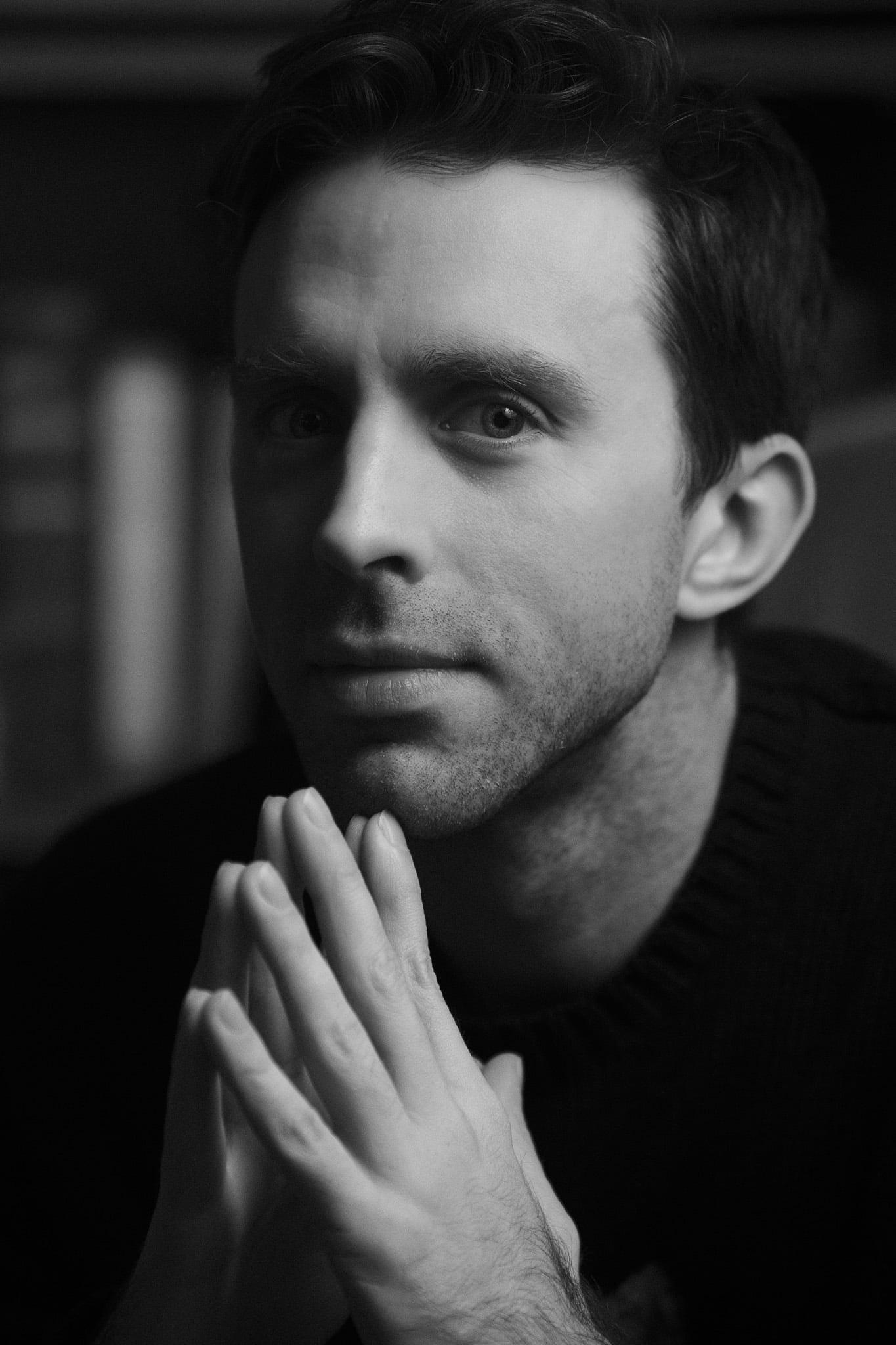 A man in a dark sweater, fingertips pressed together, in black and white.