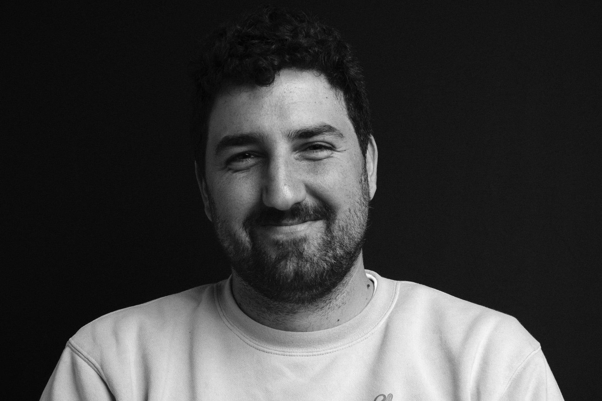 The man alone, smiling, in a white sweatshirt against a black backdrop.