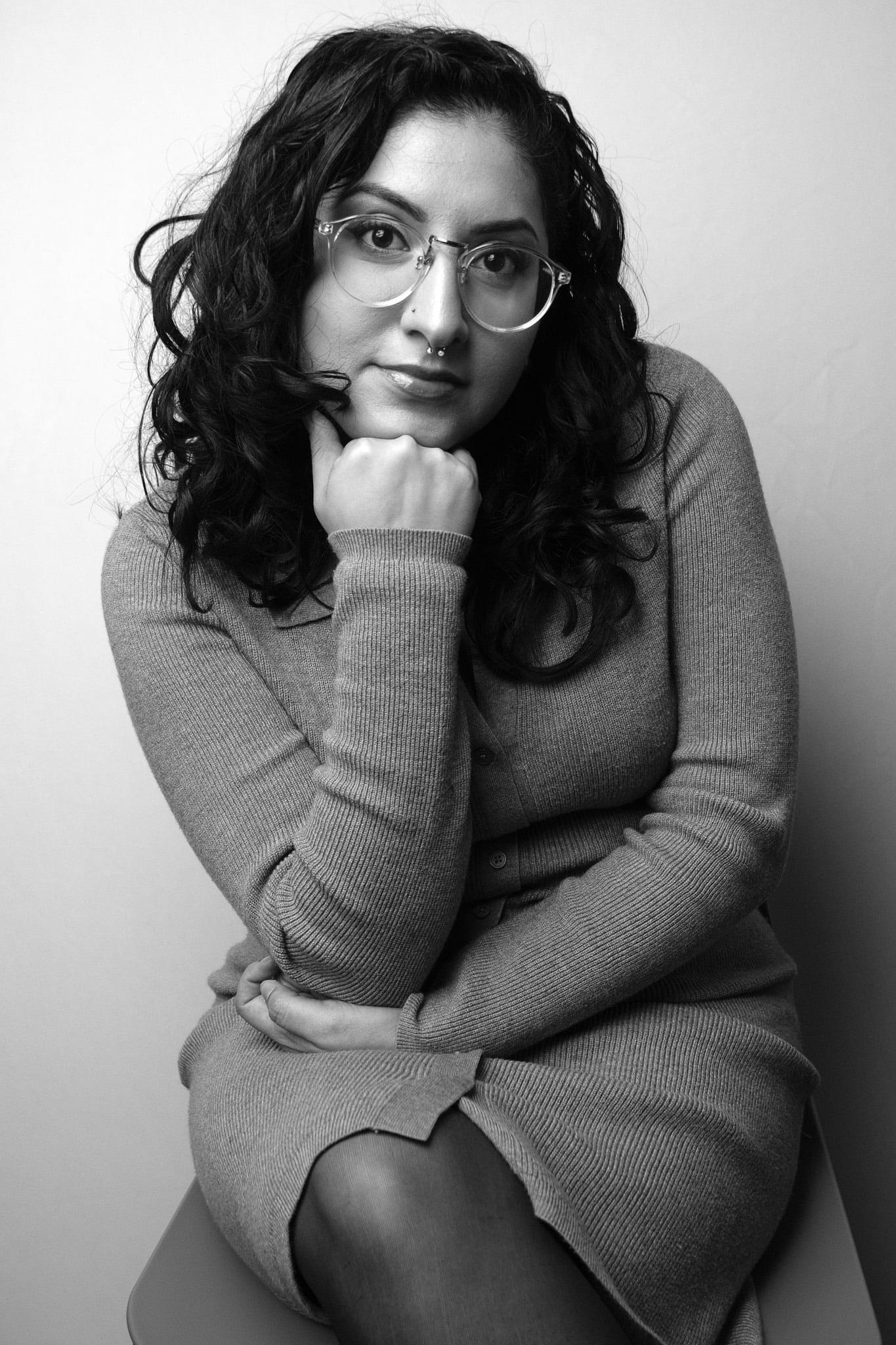 Woman with long curly hair and round glasses, seated, chin on her hand, in black and white against a light wall.