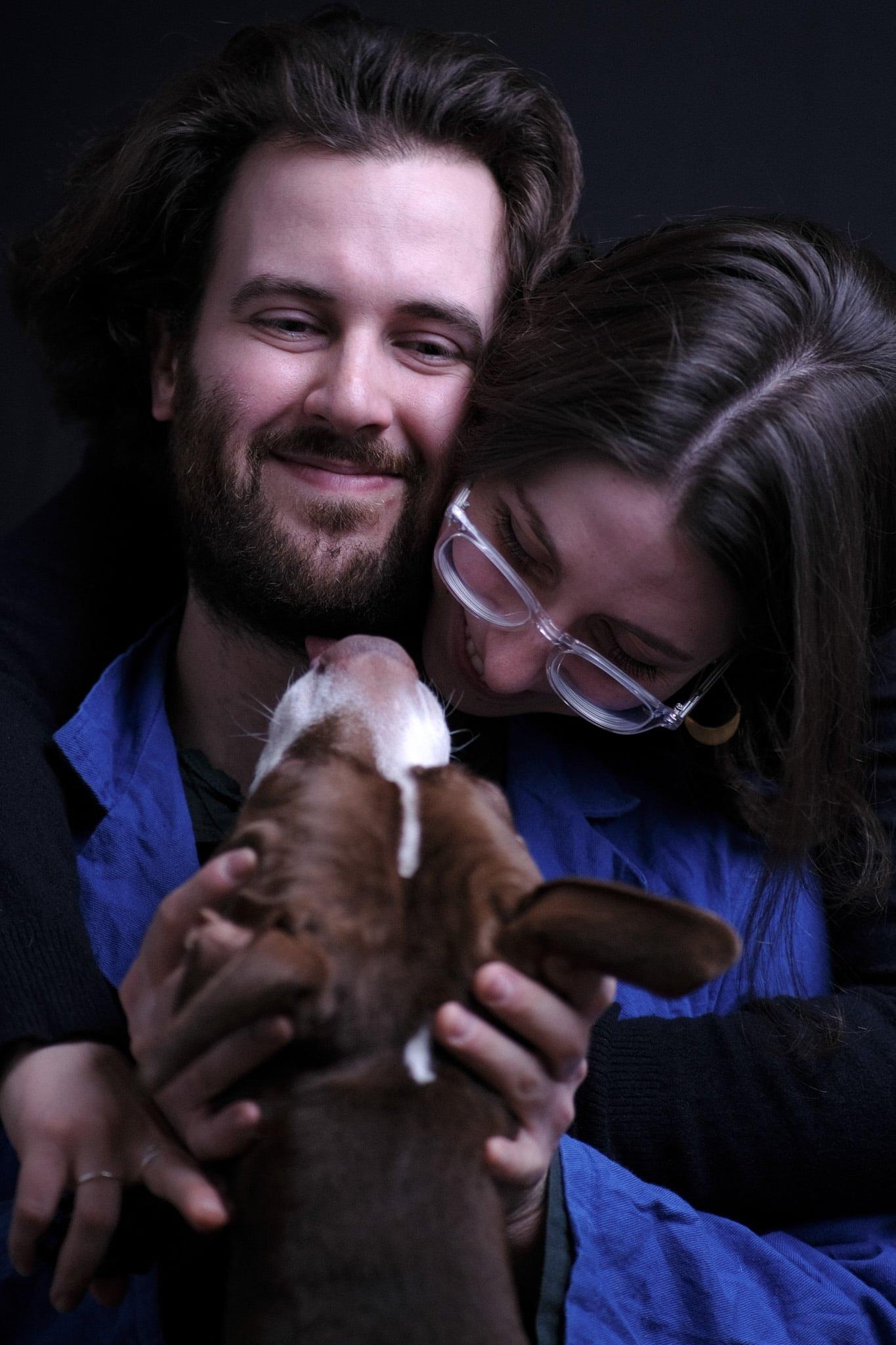 A couple hugging a dog that is licking the man’s face, lit in blue.