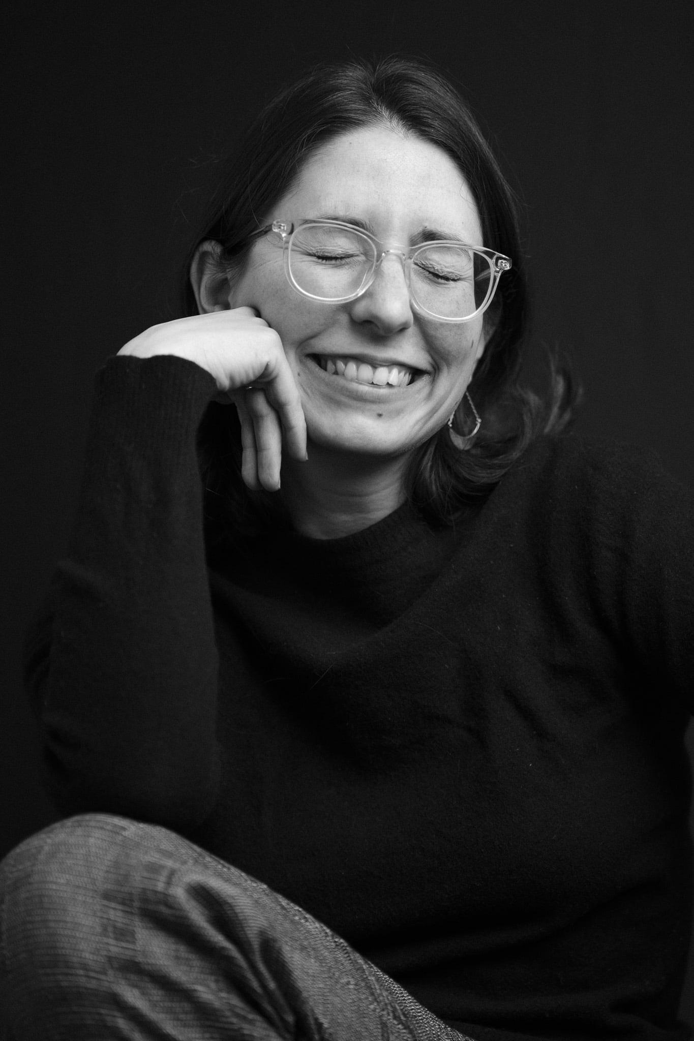 A woman in round glasses laughing, chin in her hand, in black and white.
