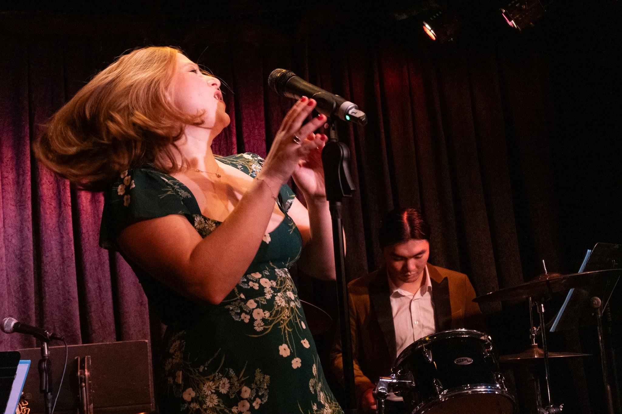 A singer throwing her head back mid-note in front of a red curtain.