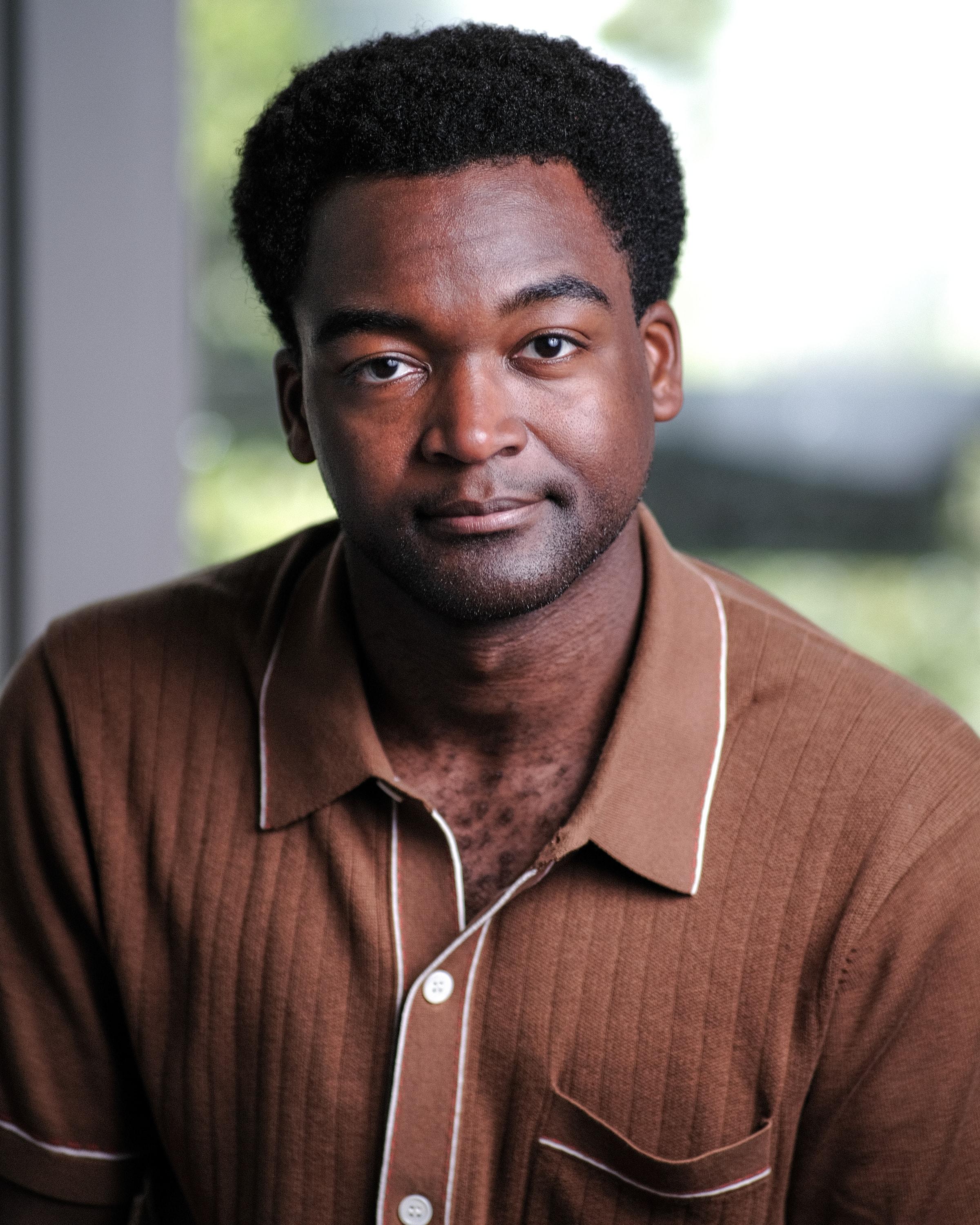 A man in a brown camp-collar shirt outdoors, green foliage blurred behind him.