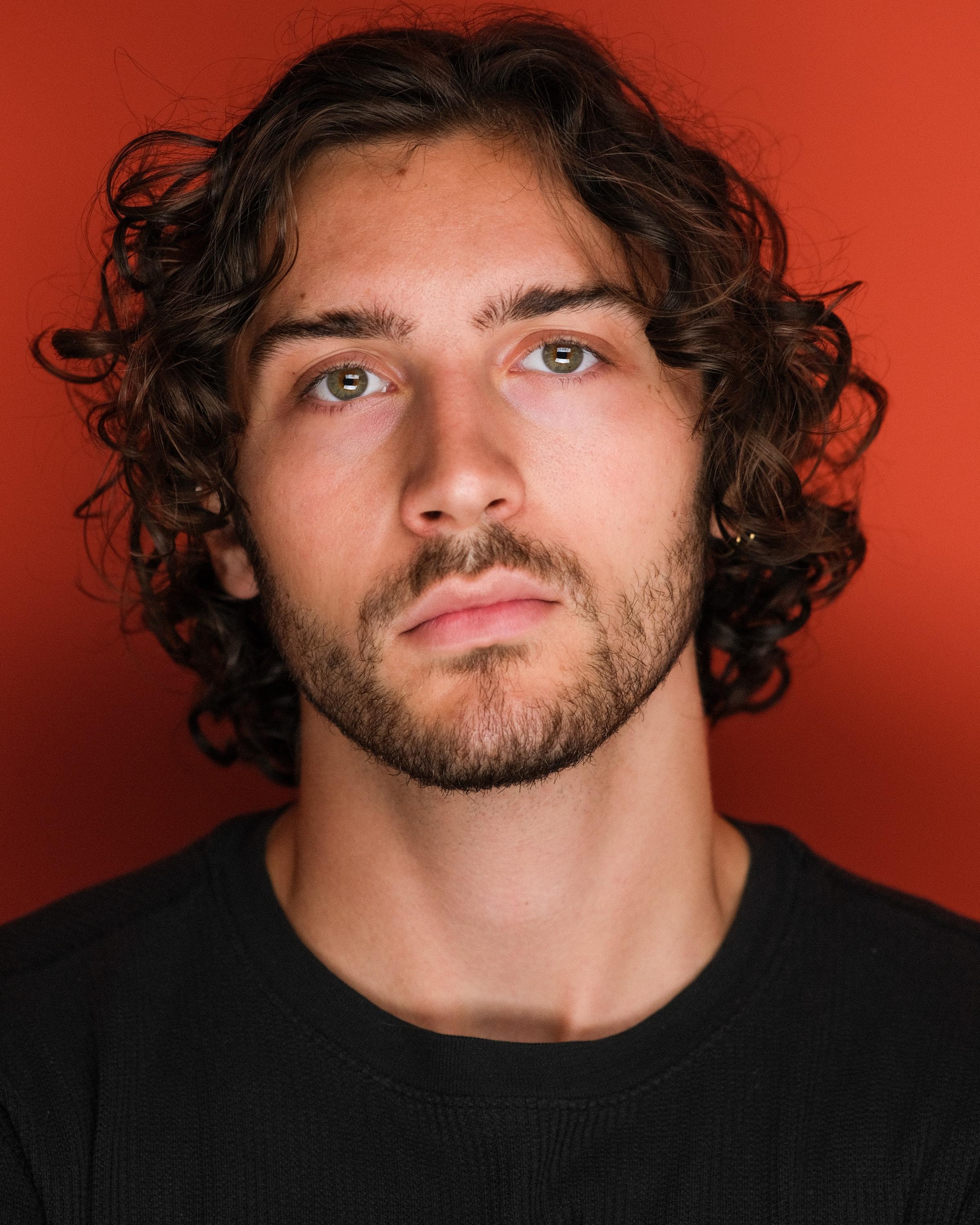 A man with shoulder-length dark curly hair in a black T-shirt against a deep red backdrop.