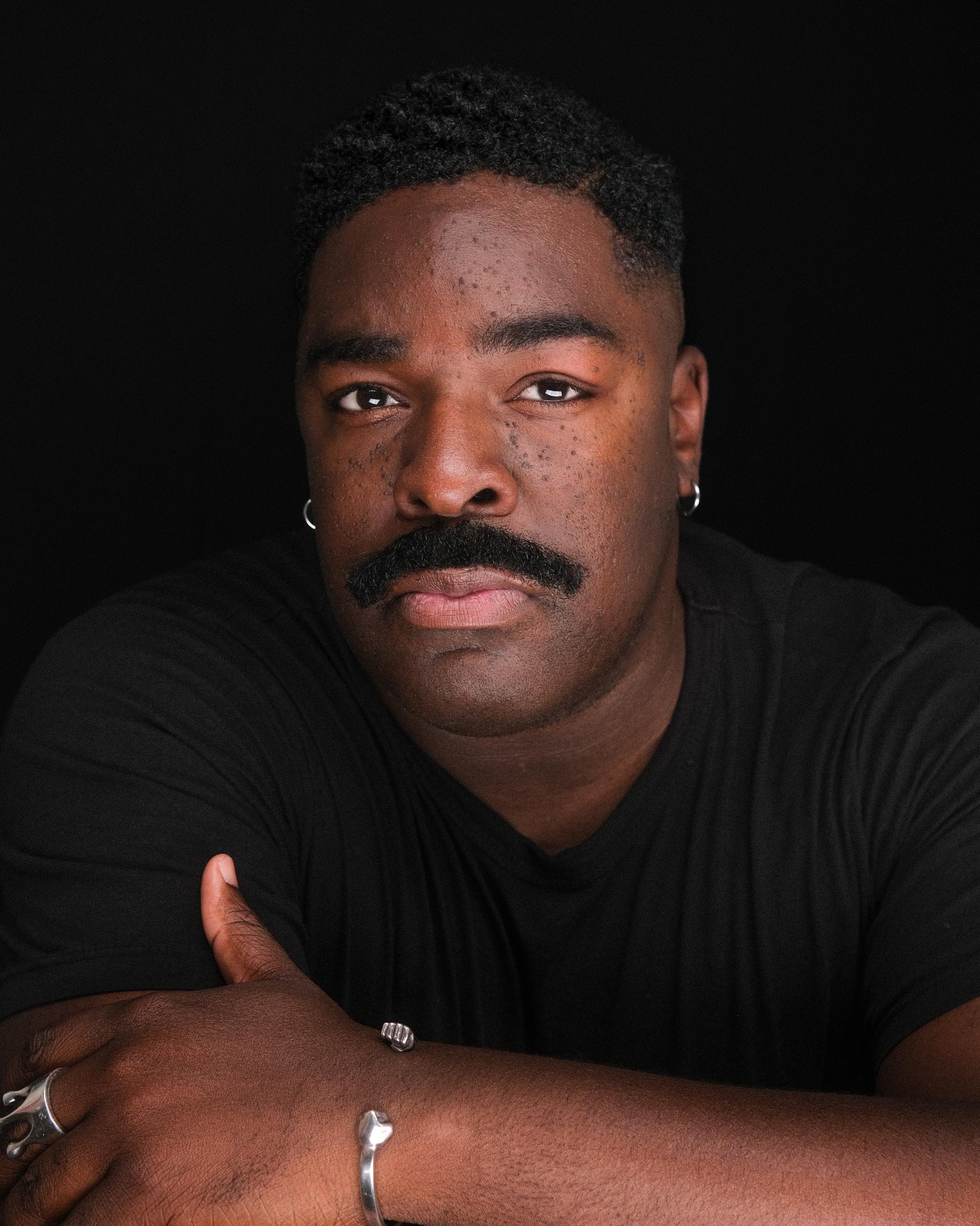 A man with a full mustache in a black T-shirt, arms folded, against a black backdrop.