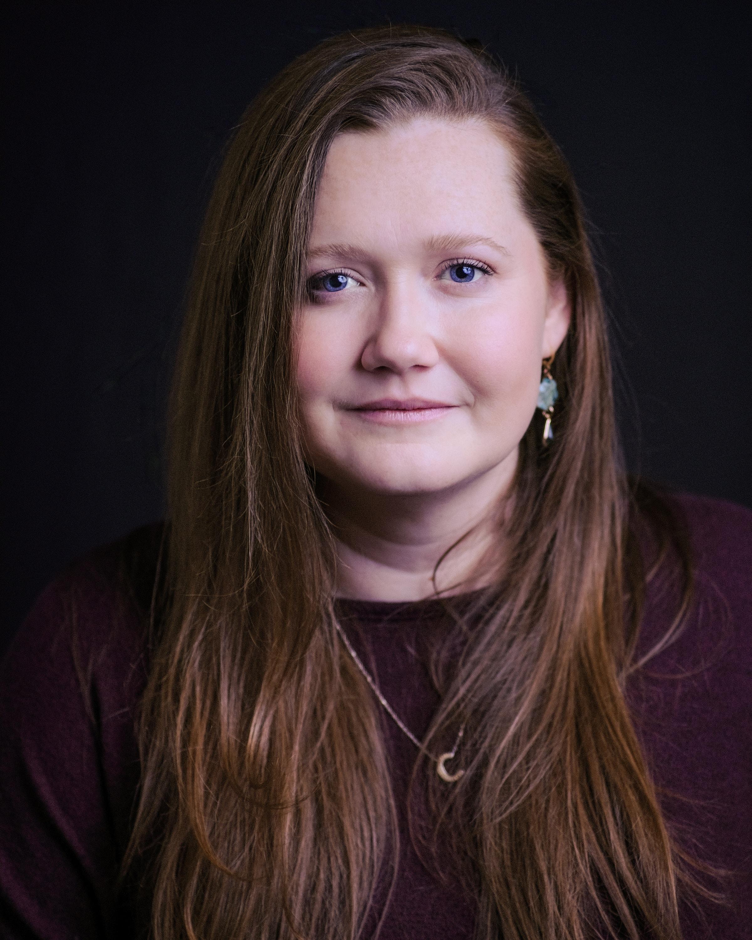 A woman with long auburn hair and a soft smile against a dark backdrop.