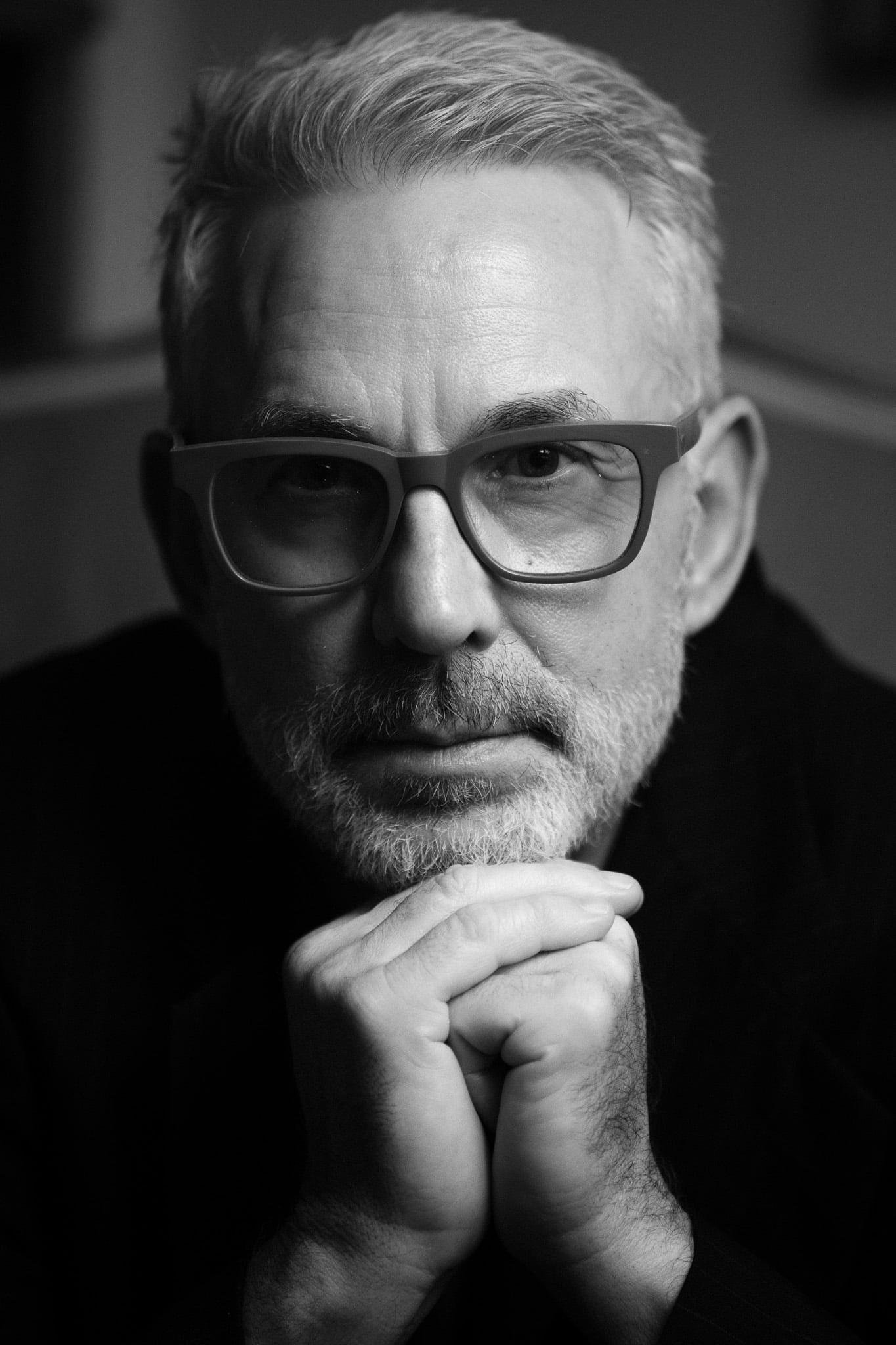 An older man in dark glasses, hands clasped, in black and white.